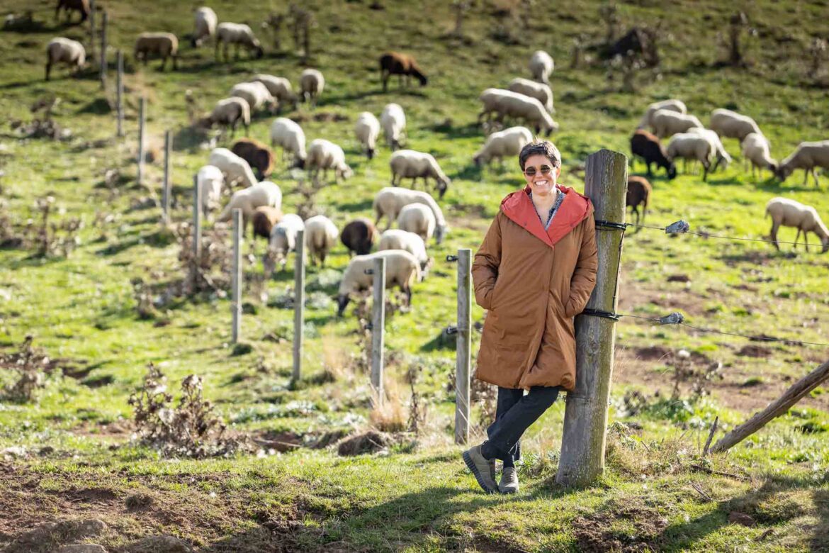 Retrato de cuerpo entero de Núria Correig en Bruguera, Girona, apoyada en una valla de montaña con un rebaño de ovejas en segundo plano para su sesión de fotografía de marca personal